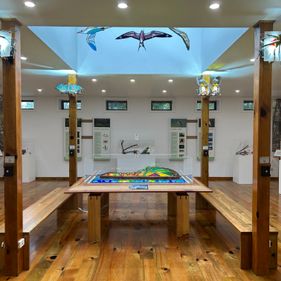 Inside the Discovery Centre, a timber strip floor and white walls. In the centre of the room is a wooden table with a stained glass sculpture in the centre. On wooden pillars are sculptures of flying animals made of stained glass and metal